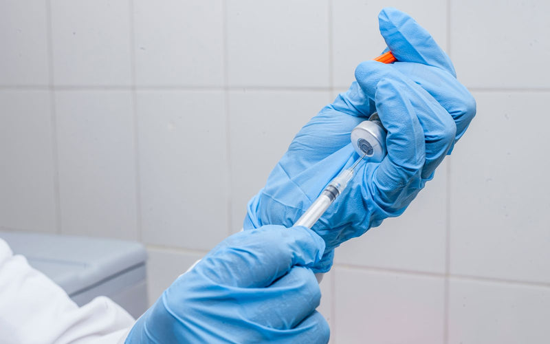 Handen met blauwe handschoenen trekken met een spuit vloeistof uit een flacon tijdens reconstitutie.