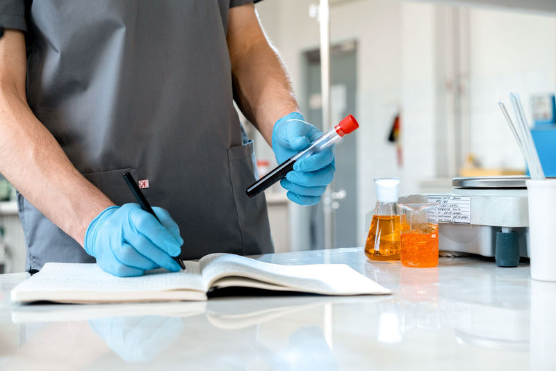 Onderzoeker met handschoenen noteert meetgegevens in een labnotitieboek naast een reageerbuis.